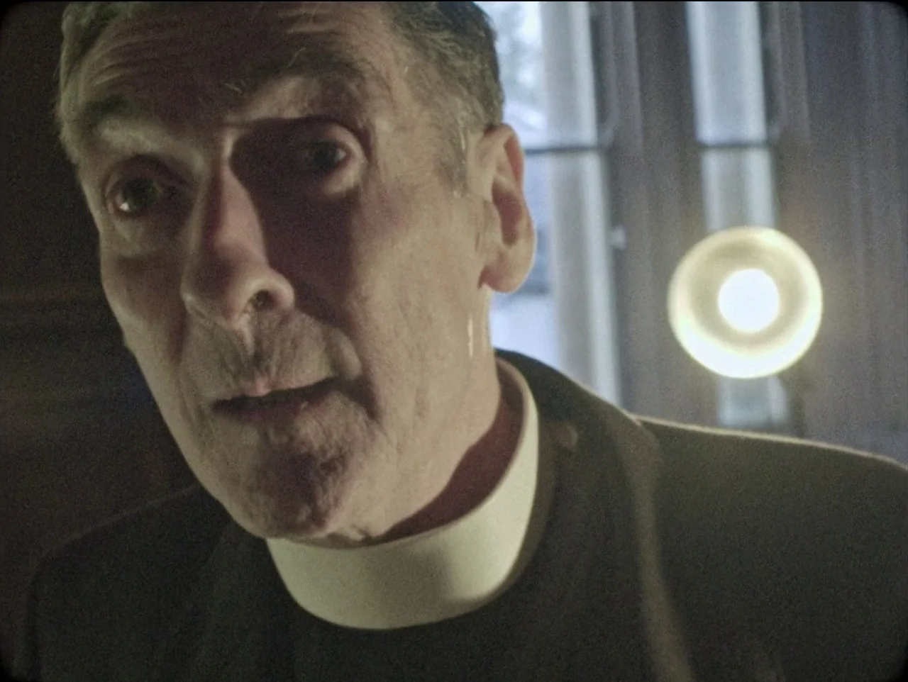 The Devil’s Doorway (2018) movie still: closeup — A dimly lit interior space contains a middle-aged man in clerical clothing. The man looks directly…