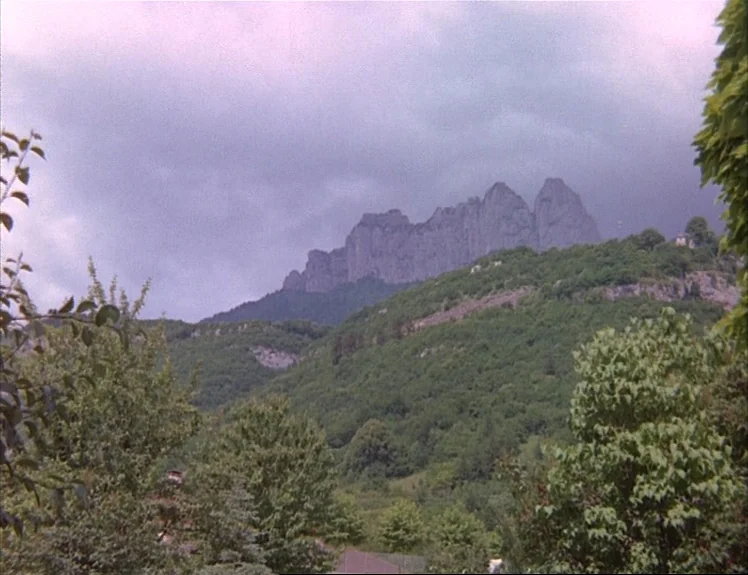 Claire’s Knee (1970) movie still: establishing — A rugged mountain range rises behind dense, rolling green hills under an overcast sky. The…