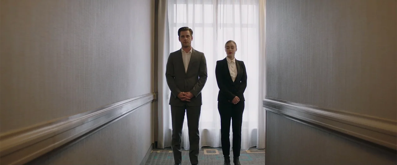 Playing God (2021) movie still: fullbody — A narrow hotel corridor with grey walls leads to a large window with white curtains. A man and a woman in…