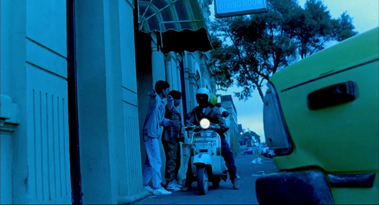 Romper Stomper (1992) movie still: wide — A blue-toned sidewalk exterior features a classic building facade with a 'Dining Room' sign. Three young…