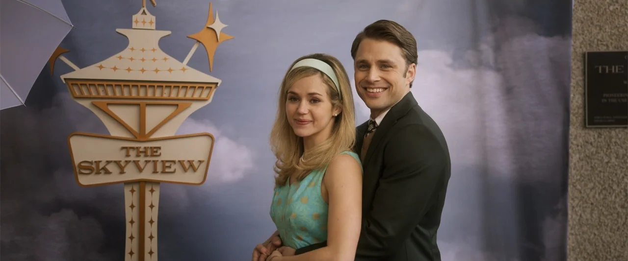 Final Destination Bloodlines (2025) movie still: medium — A man and a woman pose in front of a printed banner depicting a stylized tower. The couple…