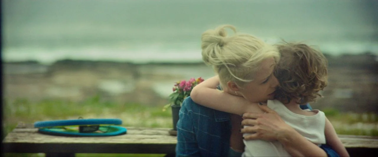 Calm With Horses (2019) movie still: medium — A wooden picnic table sits outdoors against an out-of-focus coastline backdrop. A blonde woman hugs a…