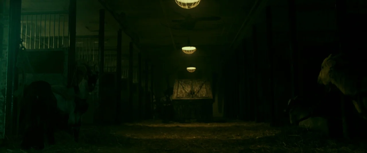 Ready or Not (2019) movie still: wide — A dark indoor barn walkway with wooden stalls on both sides and central overhead lighting. Two goats occupy…