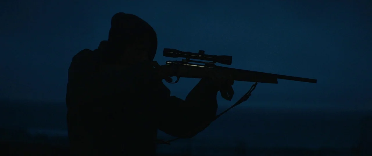 Beast (2017) movie still: medium — A dark outdoor space during the night with a faint horizon line. A person in a hooded jacket aims a scoped rifle…