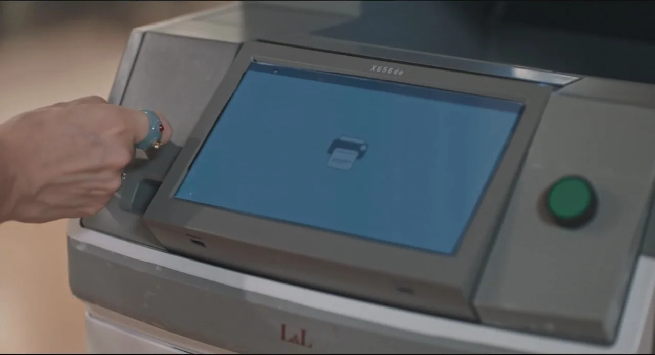 Proof of Love (2022) movie still: closeup — An office printer features a touch-screen display showing a printer icon. A hand with a teal and red…