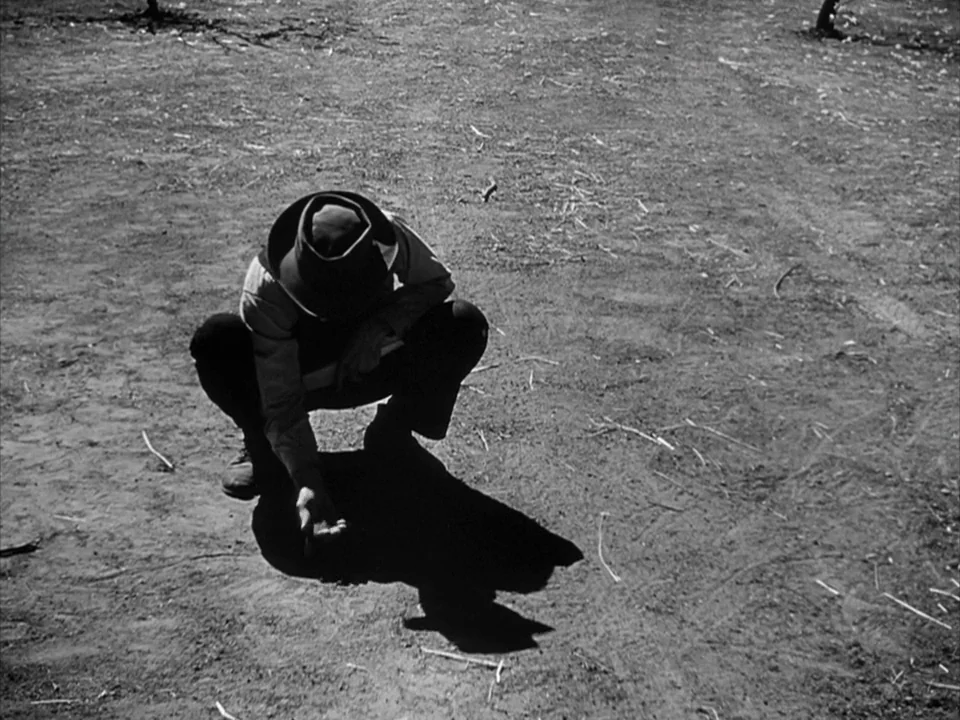 The Grapes of Wrath (1940) movie still: fullbody — A man crouches in a dry, sparse patch of dirt under bright daylight. He extends his hand toward…