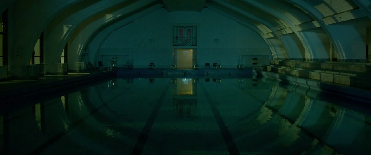 The Lure (2015) movie still: establishing — An empty indoor swimming pool features high arched ceilings and tiered concrete seating areas. The water…