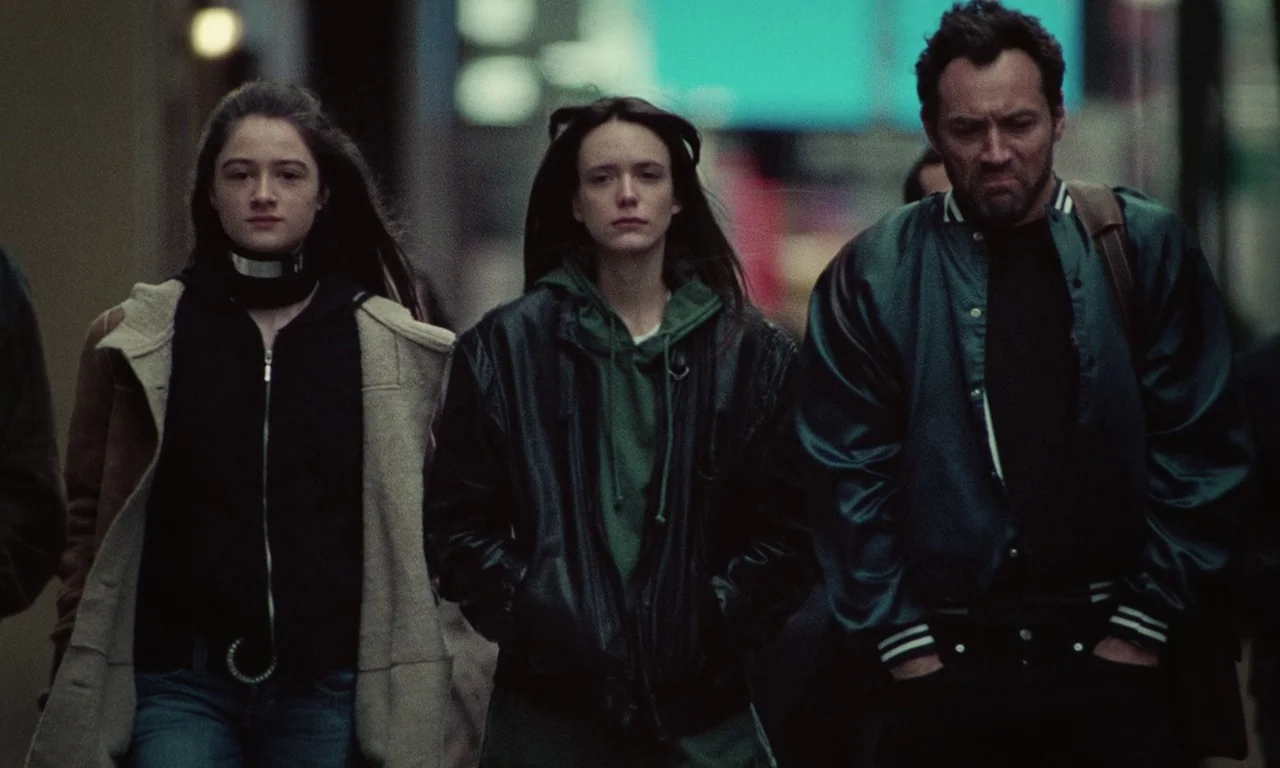 Vox Lux (2018) movie still: medium — Three people walk closely together along a dim, out-of-focus urban sidewalk at night. The individuals move…