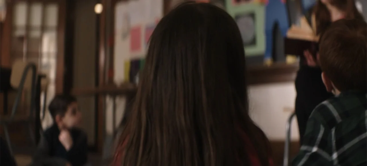 I Still See You (2018) movie still: medium — A classroom interior with children sitting in a cluster listening to a speaker. A girl with dark hair…