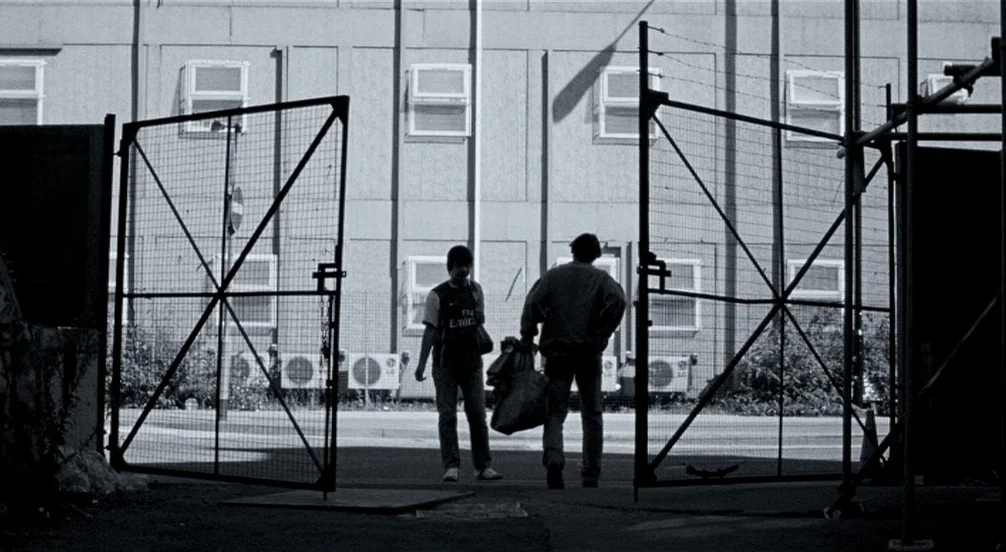 Somers Town (2008) movie still: wide — Two individuals stand in an industrial opening framed by large metal gates and fences. A concrete building…