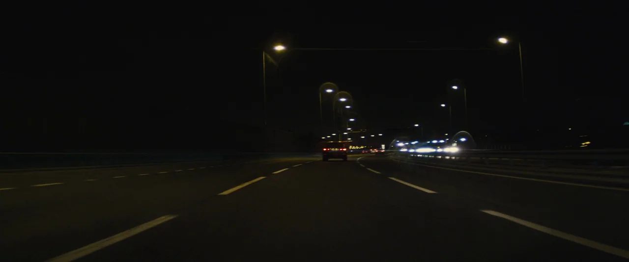 Locke (2013) movie still: wide — A multi-lane highway stretches into the dark distance under artificial overhead street lighting. Several vehicles…
