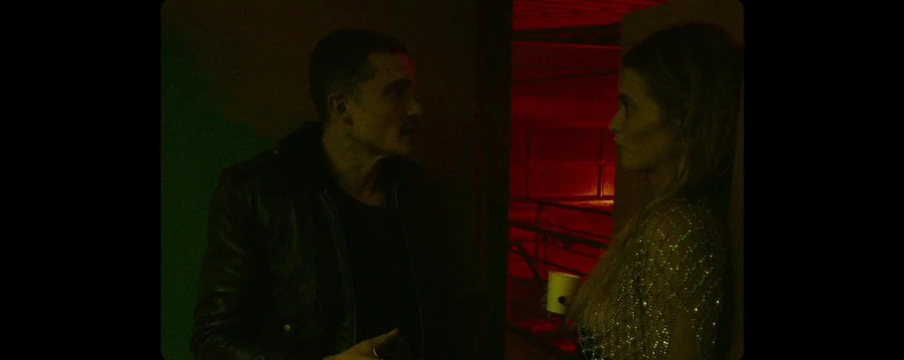 Lux Æterna (2019) movie still: medium — A dark, cramped interior space is illuminated by vibrant red and green neon lights. A man in a leather…
