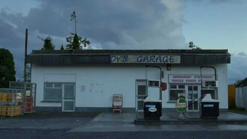 Garage (2007) movie still: wide — A quiet, weathered gas station with a white facade sits under a cloudy sky. The building features old signage and…