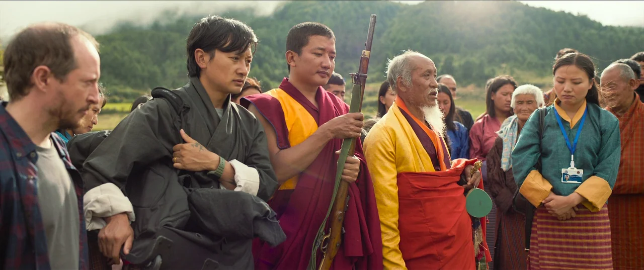 The Monk and The Gun (2023) movie still: medium — A diverse group of people stands on a grassy mountain path during the day. A monk holds a vintage…