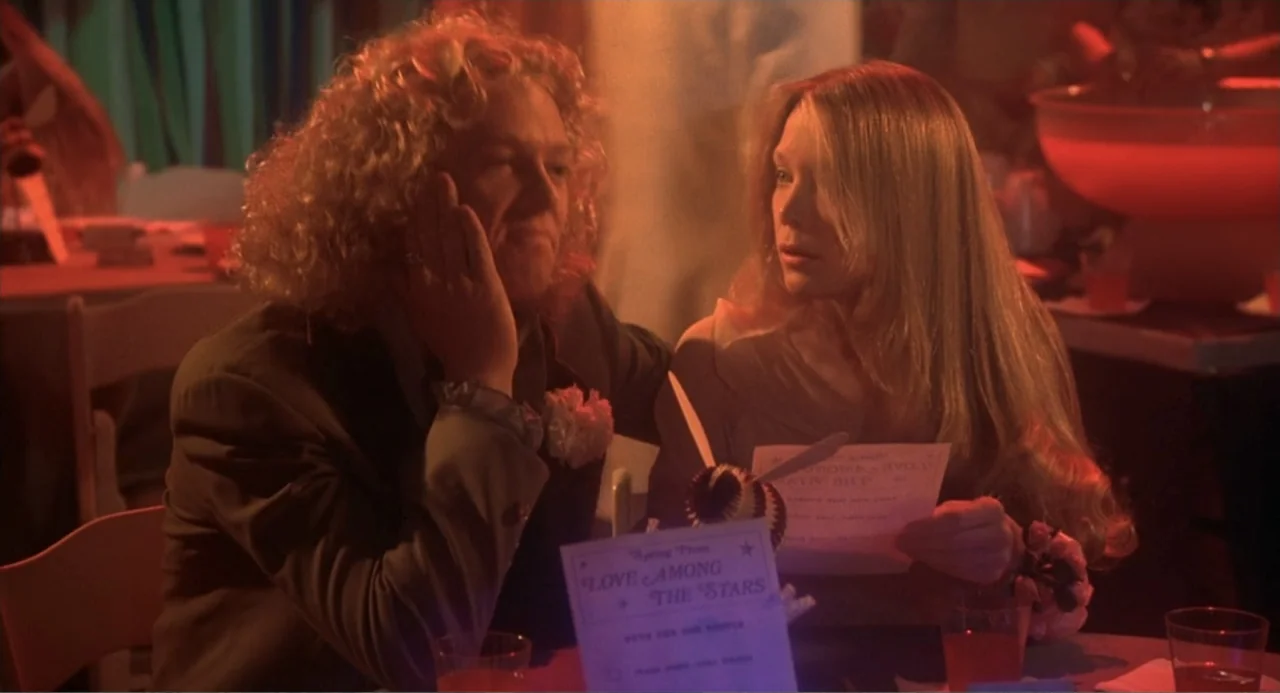 Carrie (1976) movie still: medium — A high school gym is decorated for a prom dance with red lighting. A young man and woman sit at a table reading…
