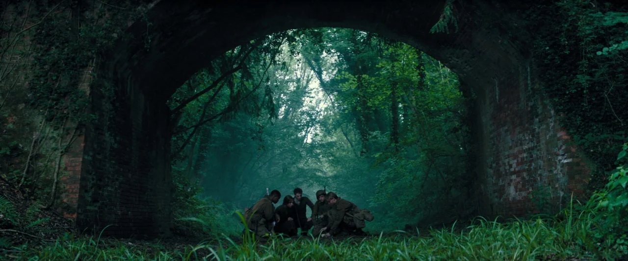 Overlord (2018) movie still: wide — A group of five soldiers crouches under an ivy-covered brick archway in a dense forest. The men huddle together…