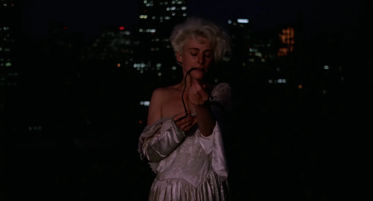 Liquid Sky (1982) movie still: medium — A woman stands on a rooftop against a dark, out-of-focus city skyline at night. She holds a thin, dark cable…
