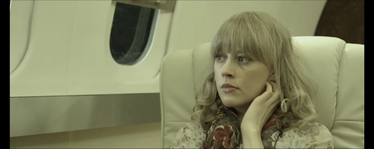 Turbulence (2025) movie still: closeup — A woman sits in a cream-colored leather seat within an aircraft cabin. She rests her hand against her head…