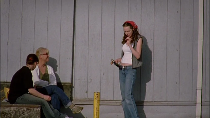 Old Joy (2006) movie still: fullbody — A group of three women stands against a weathered grey vertical wood plank wall. The standing woman holds a…