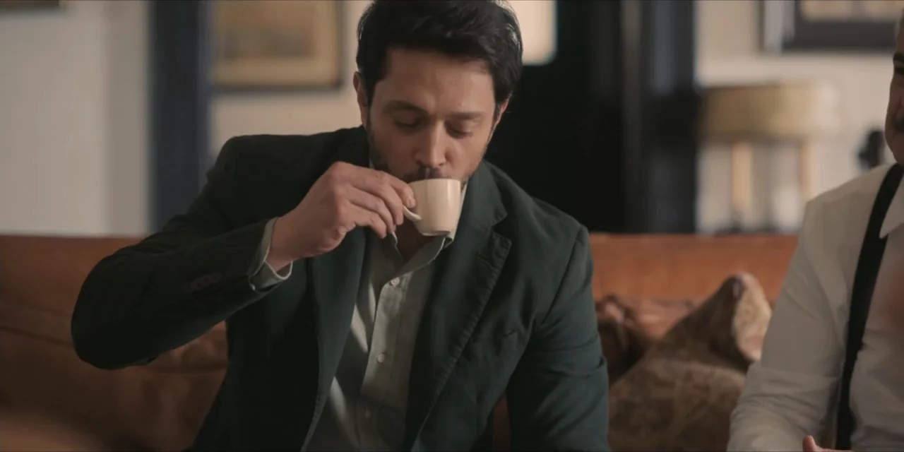 Kill Me If You Dare (2019) movie still: medium — An interior living room with a brown leather sofa and blurred background decor. A man in a dark…