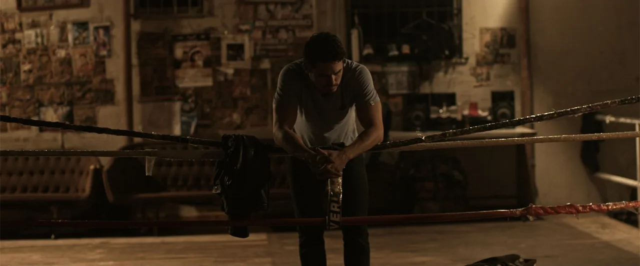 Kimura (2017) movie still: medium — A dimly lit room contains boxing ropes and posters covering the back wall. A man leans forward on the ropes with…