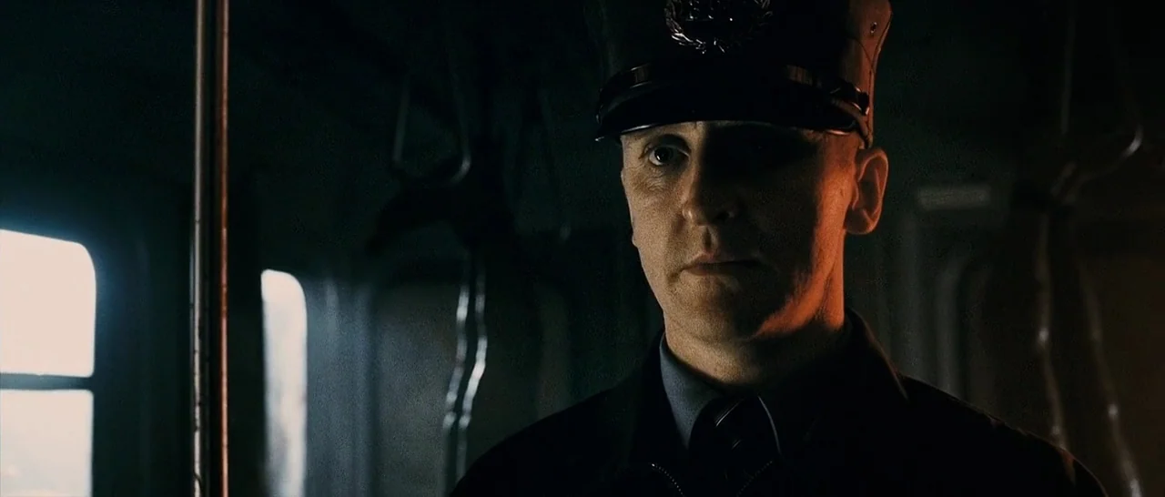The Midnight Meat Train (2008) movie still: closeup — A dimly lit subway car interior has metallic poles and dark window frames. A man in a formal…
