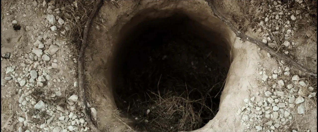Matar o morir (1984) movie still: closeup — A deep circular hole is dug into dry, light-colored dirt and rocky terrain. The interior of the hole is…