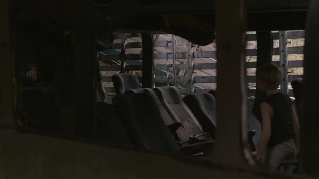 Boy (2010) movie still: medium — The interior of a derelict bus with worn fabric seats and exposed frame structural members. A young child with…