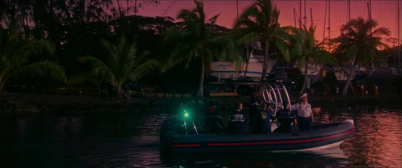Pacifiction (2022) movie still: wide — A rigid-hulled inflatable boat travels along a shoreline lined with palm trees at dusk. Several individuals…