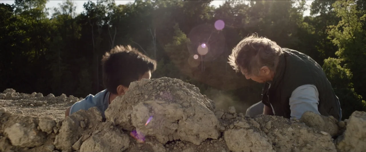 The Marksman (2021) movie still: medium — A dusty outdoor clearing features a large heap of earth in the foreground. A child and an older man work…