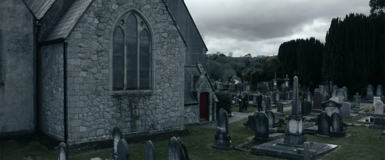 Freud's Last Session (2023) movie still: wide — A grey stone church with a red door sits adjacent to a grassy cemetery. Several men in overcoats…