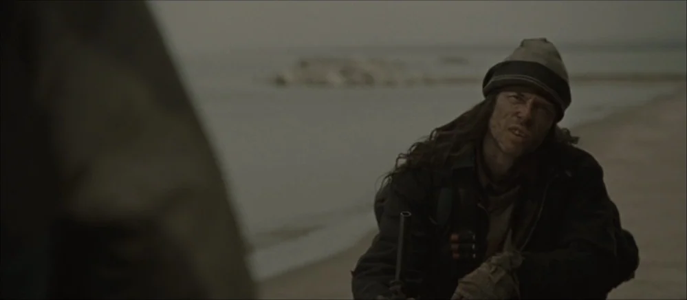 The Road (2009) movie still: medium — An overcast beach setting with gray water and sand stretching toward a distant coastline. A man in a knit cap…