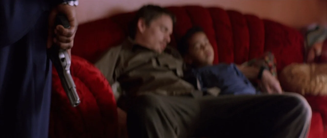 Training Day (2001) movie still: medium — A man and a child are sleeping on a red curved sofa. In the foreground, an out-of-focus hand holds a…
