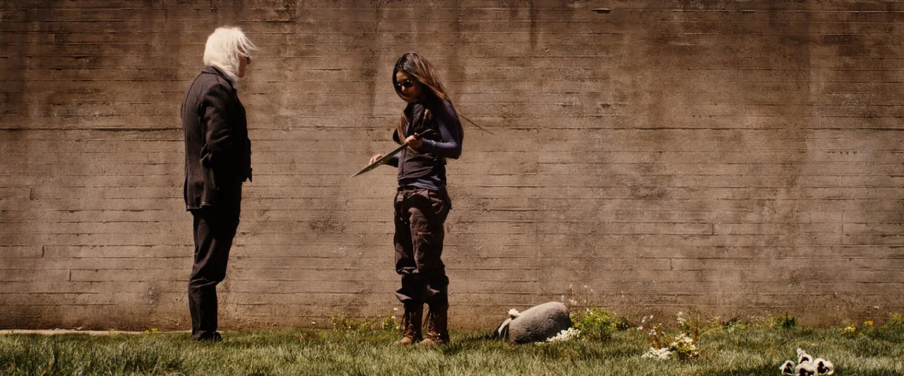 The Book of Eli (2010) movie still: fullbody — Two individuals stand in a grass field before a textured concrete wall. A young woman examines a…