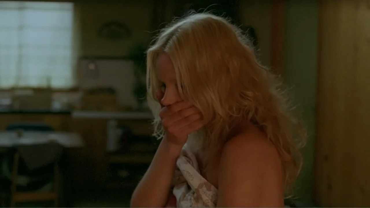Emily (1976) movie still: closeup — A dimly lit room features a blurred background with a window and wooden surfaces. A blonde woman in her twenties…
