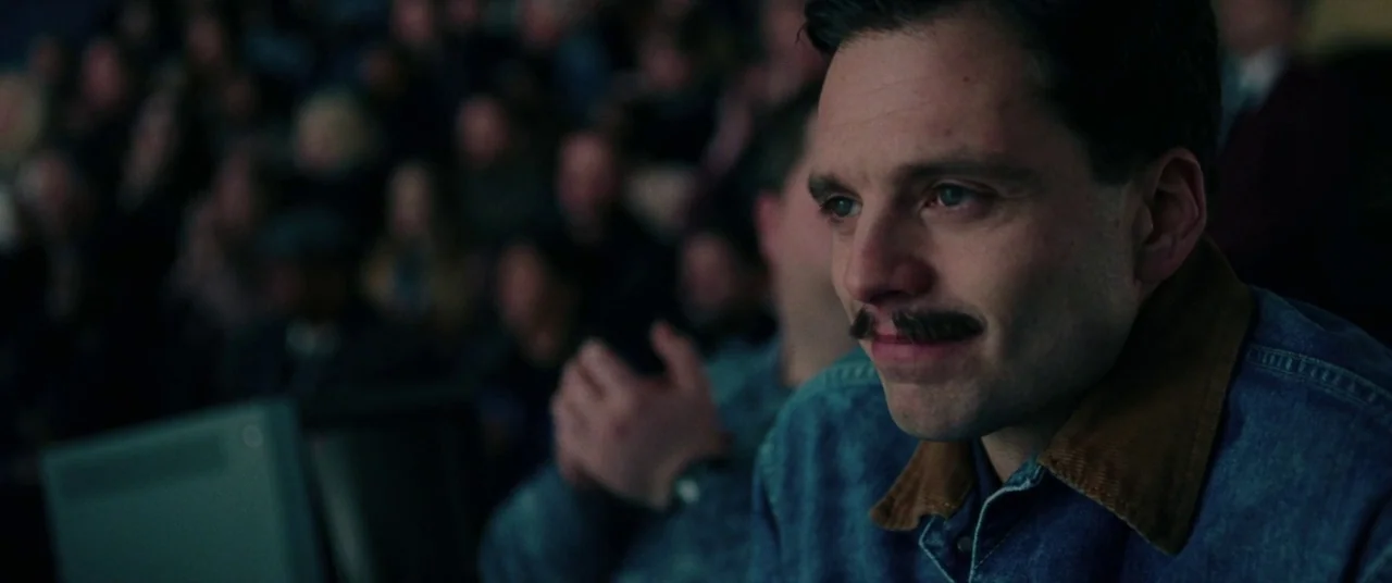 I, Tonya (2017) movie still: medium — A dimly lit auditorium is filled with a blurred audience watching from their seats. A man with a mustache sits…
