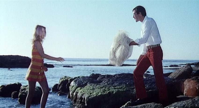 5 Dolls for an August Moon (1970) movie still: fullbody — A rocky shoreline with mossy stones meets the shallow edge of the sea. A man in a white…
