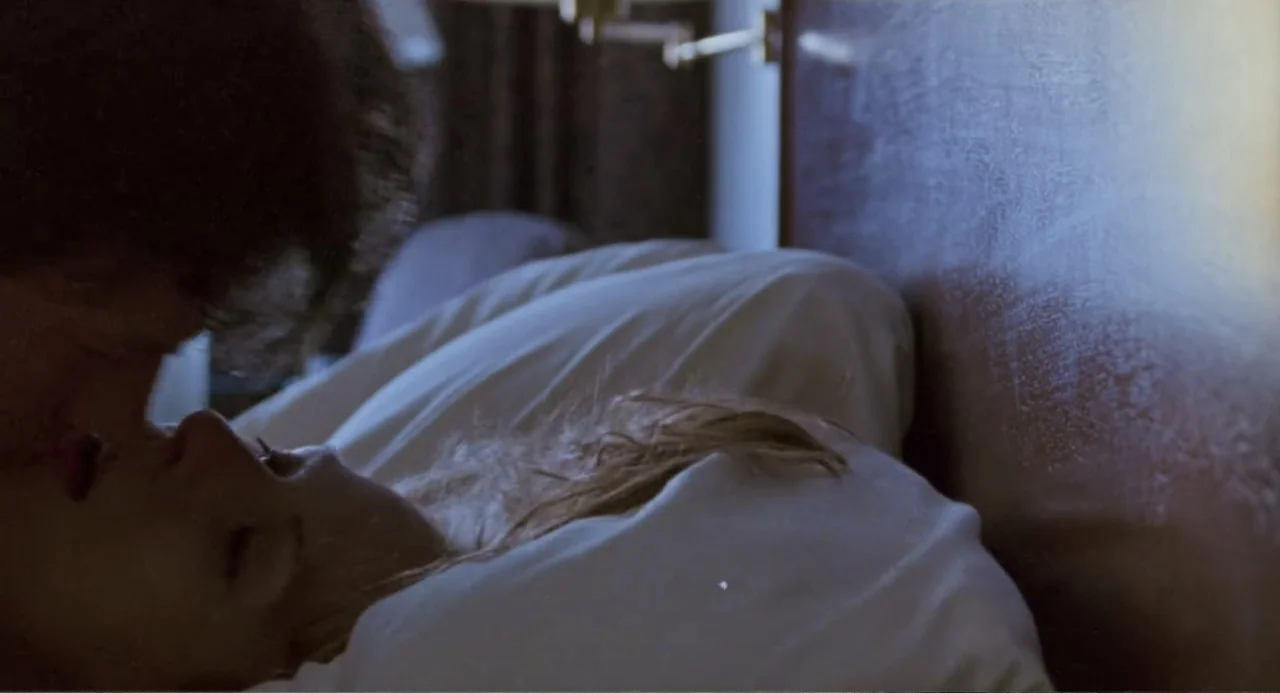 Forty Shades of Blue (2005) movie still: closeup — Two individuals are lying in a bed with white sheets and a dark headboard. A man and a woman are…