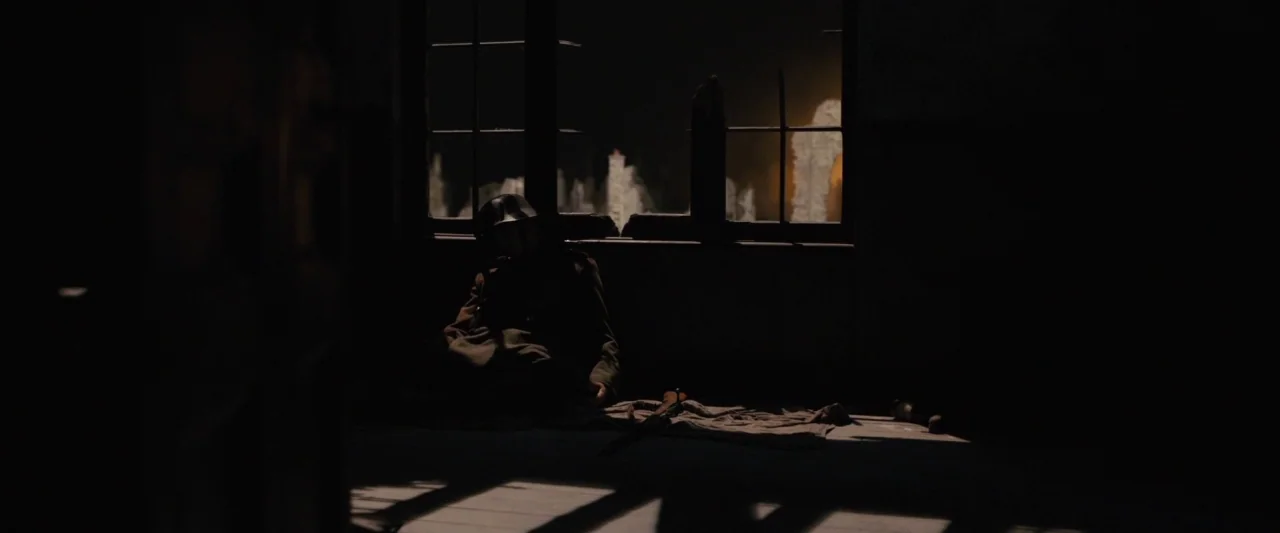 1917 movie still: wide — A dark, dilapidated interior space features a single window looking out toward distant city ruins. A soldier wearing a…