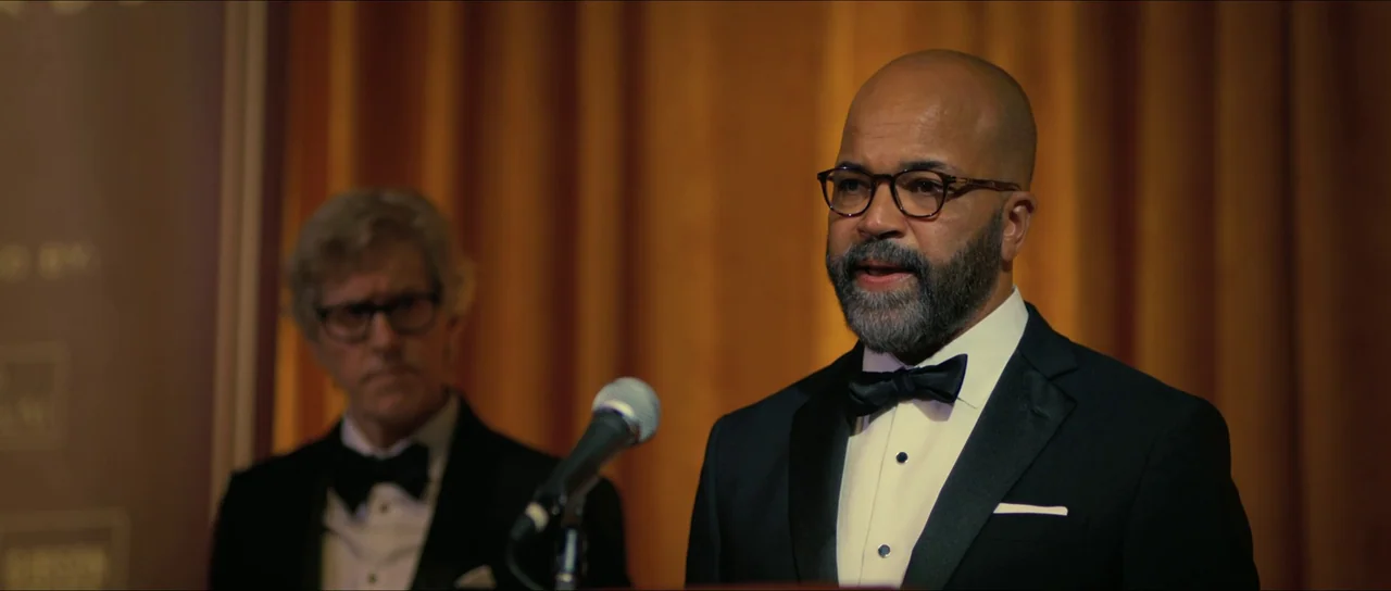 American Fiction (2023) movie still: medium — A man in a tuxedo stands at a podium in front of gold curtains. Another man in a tuxedo stands…