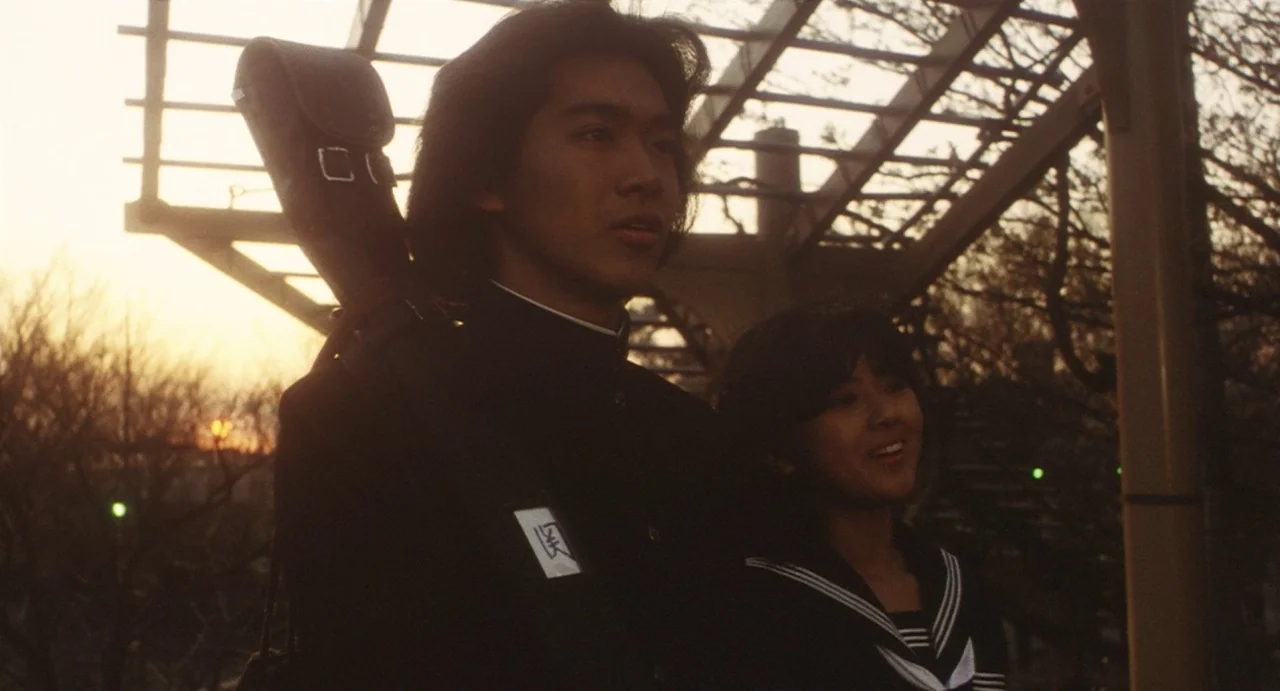 School in the Crosshairs (1981) movie still: medium — An outdoor train station platform features an overhead metal structure during sunset. A male…
