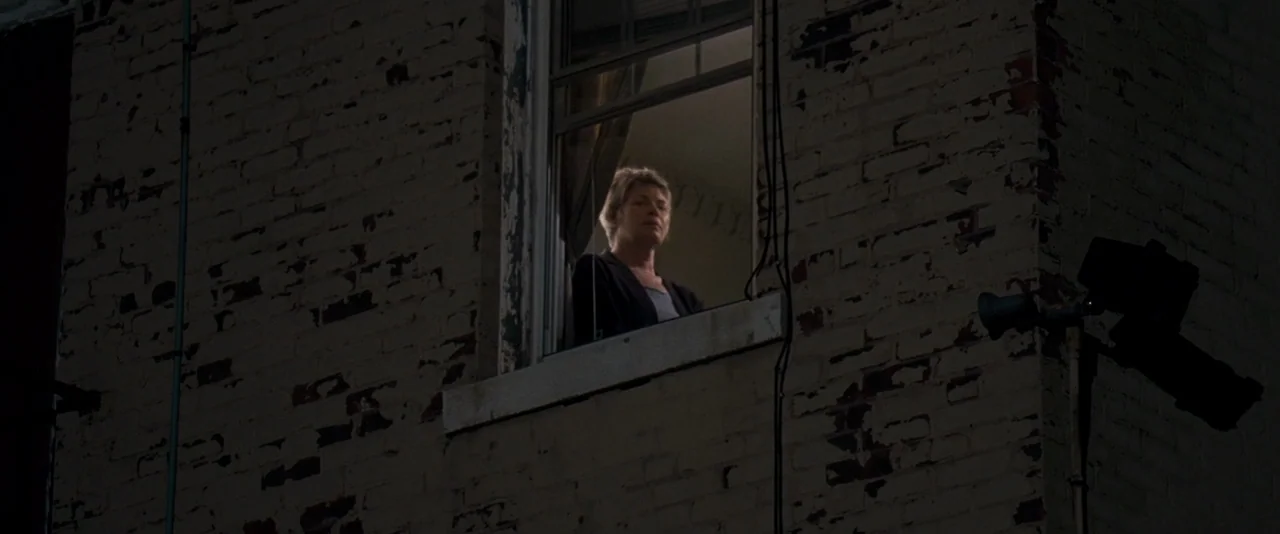 The Innkeepers (2011) movie still: medium — A weathered brick wall at night features a single window with a visible occupant. The woman looks down…