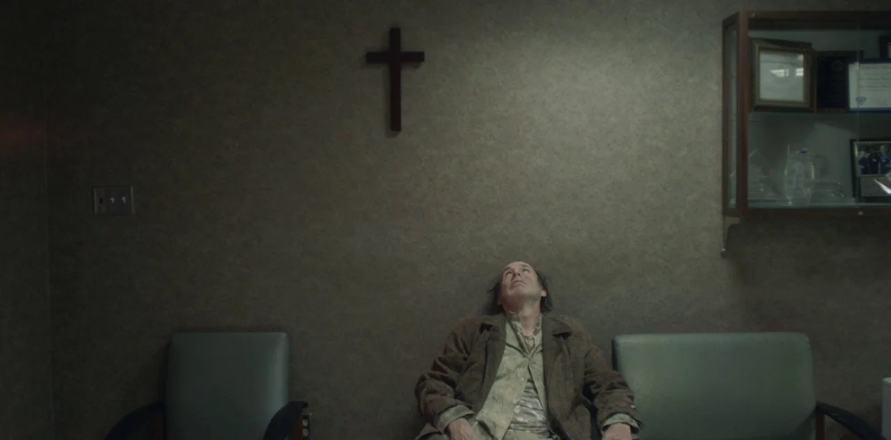 Darlin’ (2019) movie still: medium — A spartan waiting room features a textured grey wall decorated with a wooden cross. A middle-aged man sits…