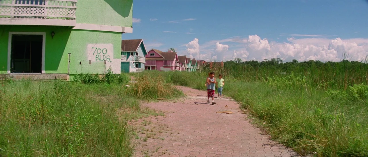 The Florida Project (2017) movie still: wide — A row of brightly painted pastel houses stands beside an overgrown marshy field. Two young children…