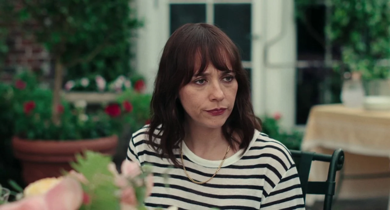 On The Rocks (2020) movie still: medium — A woman sits at an outdoor table with a brick building and plants behind her. She looks to the side while…
