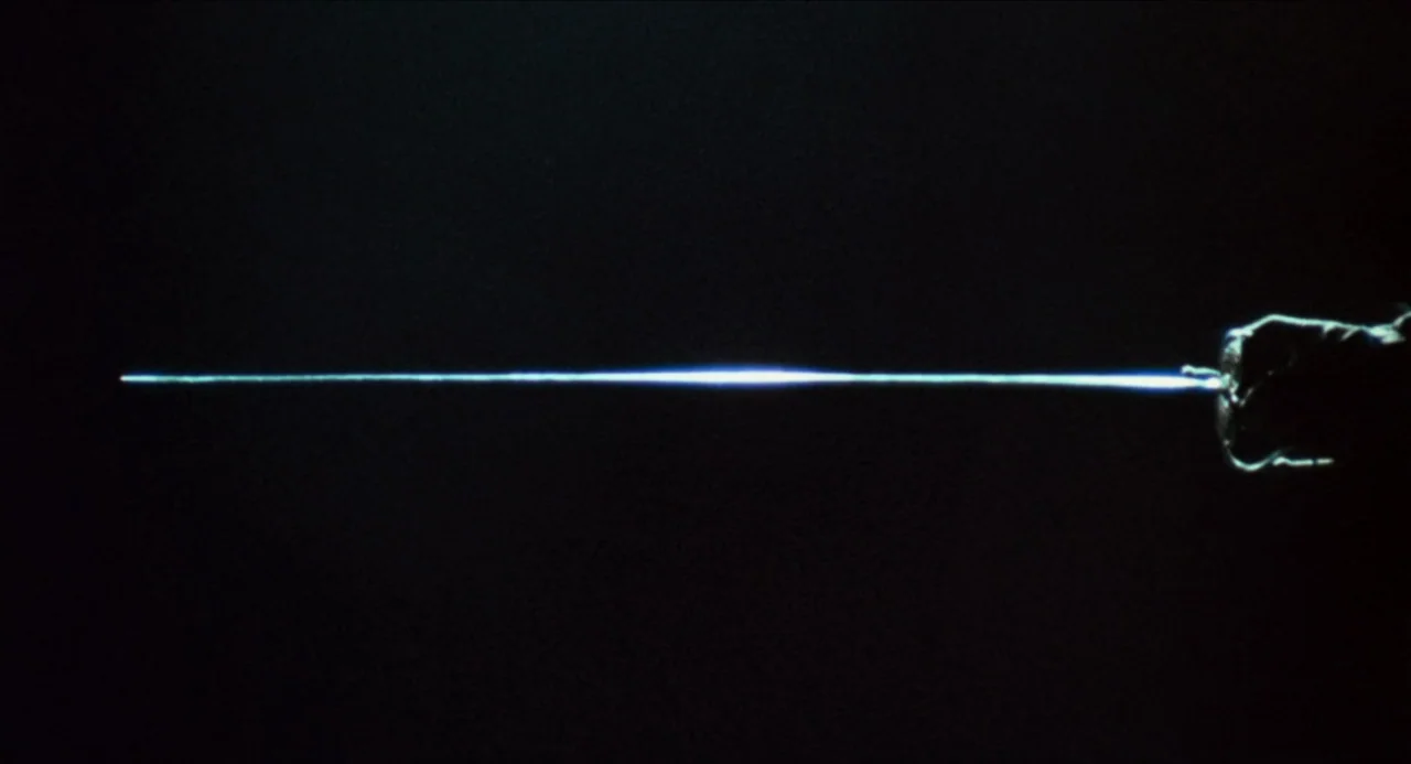 The Three Musketeers (1973) movie still: closeup — A horizontal, vibrant blue beam of light emanates from a partially visible dark hilt. The rest of…