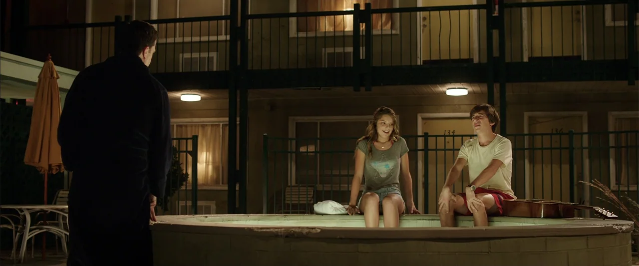 Vacation (2015) movie still: medium — A two-story motel courtyard at night with a small tiled pool in the foreground. A teenage boy and girl sit on…