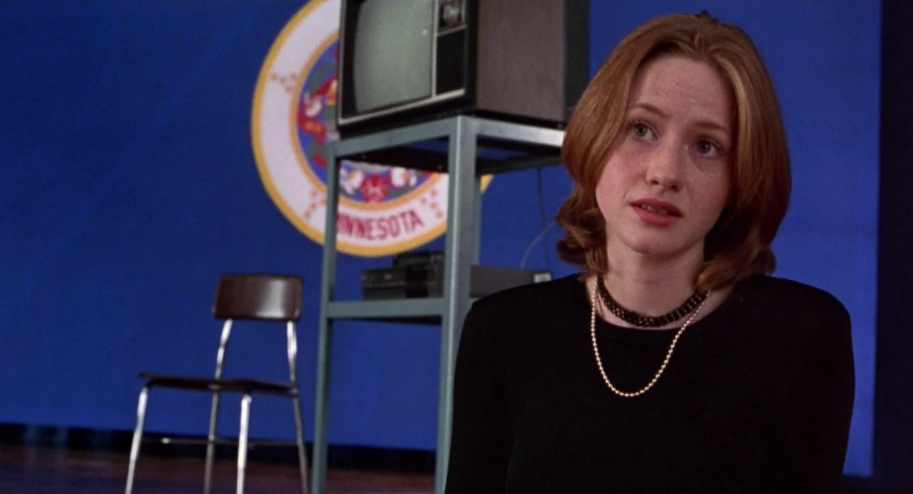 Drop Dead Gorgeous (1999) movie still: medium — A woman sits in front of a blue wall featuring a large Minnesota seal emblem. A vintage television…
