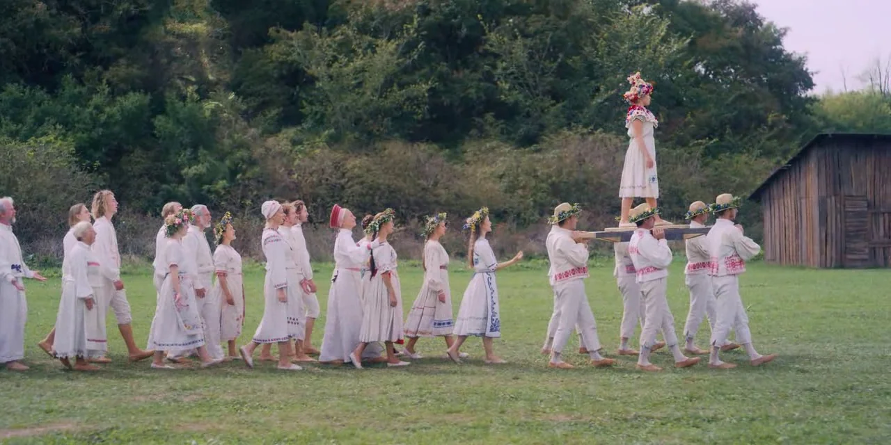 Midsommar (2019) movie still: wide — A grassy field with a wooden shed and dense treeline in the background. A group of people dressed in…