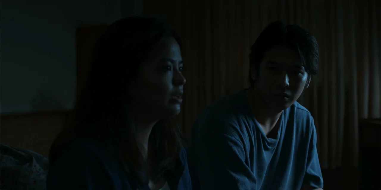 Nokturno (2024) movie still: medium — A dark, sparsely furnished interior features two people sitting together in the foreground. They remain still…